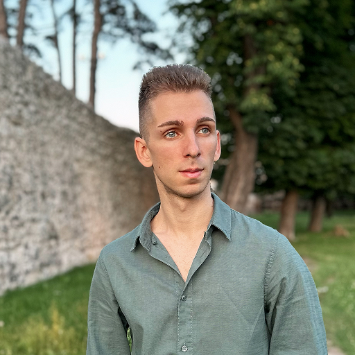 A young man with short hair wearing a green Bitcube button-up shirt stands outdoors near a stone wall and green trees, looking slightly to the side with a neutral expression.