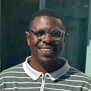 A person with short hair and glasses smiles while standing in front of a glass building. They wear a green and white striped polo shirt.