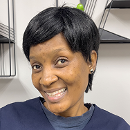 A woman with short black hair smiles at the camera. She is wearing a dark top and stud earrings, standing in front of a white wall with black geometric shelves.