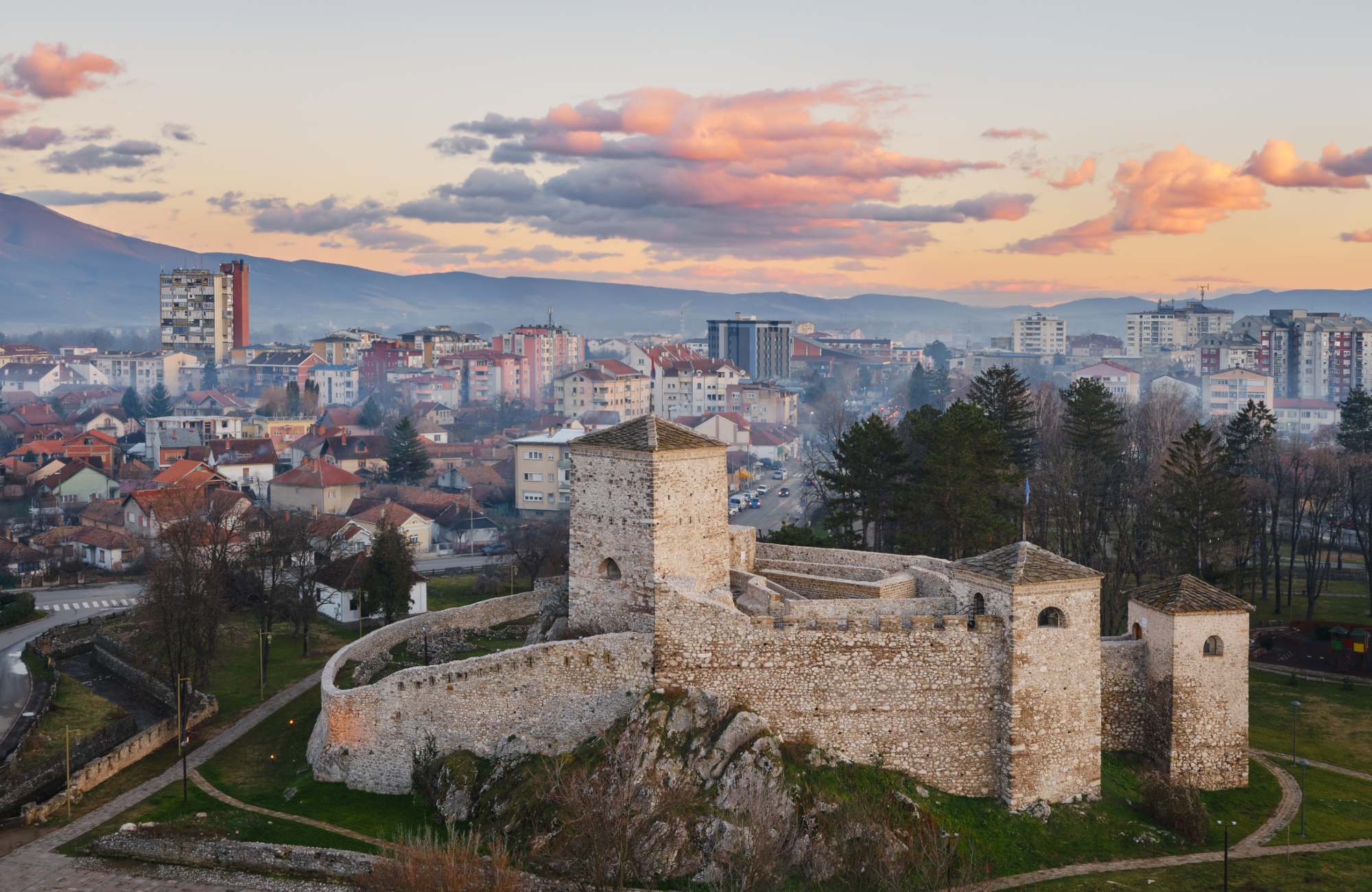 A historic stone fortress with towers and walls stands on a rocky hill, surrounded by trees and the modern Bitcube contact city skyline, all beneath a colorful sunset sky.