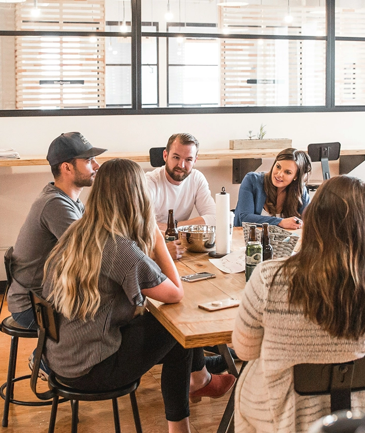 Five people sit around a wooden table in a modern office, engaged in conversation about bespoke software development. Drinks, snacks, and phones rest on the table. Large windows with blinds are in the background, setting a casual, collaborative mood.