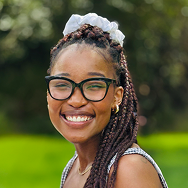 A young woman with long braids, wearing glasses and a white scrunchie, smiles outdoors with a blurred green background.