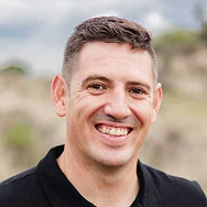 A man with short brown hair, wearing a black collared shirt, is smiling outdoors with a blurred natural background.