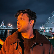 A man with dark hair and a beard stands near a waterfront at night, illuminated by orange lighting. In the background, city lights and a large ship with the word EXPLORER are visible.