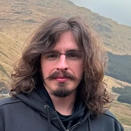 A person with long brown hair, a mustache, and a beard, wearing glasses and a black hoodie, stands outdoors with grassy hills and a cloudy sky in the background.