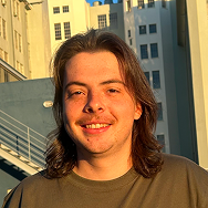 A young person with long brown hair and a light mustache smiles while standing outdoors in front of tall, light-colored buildings, with sunlight casting a warm glow on their face.