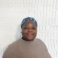 A person wearing a blue headscarf with white patterns and a beige sweater stands in front of a white brick wall, smiling softly at the camera.