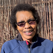 A person with short curly hair and sunglasses smiles outdoors, wearing a blue Bitcube jacket. Behind them is a rustic wooden fence in bright sunlight.