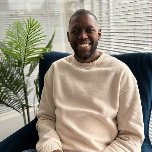 A man wearing a cream-colored sweater sits on a dark blue Bitcube chair next to a green plant, smiling, with white blinds and sunlight in the background.