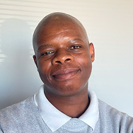 A man with a shaved head smiles softly while facing the camera. He is wearing a light gray Bitcube collared shirt and standing against a plain, light-colored background.