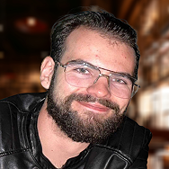 A man with a dark beard and glasses, wearing a black leather jacket, smiles at the camera. The softly blurred background with warm lighting hints at Bitcube’s inviting indoor ambiance.