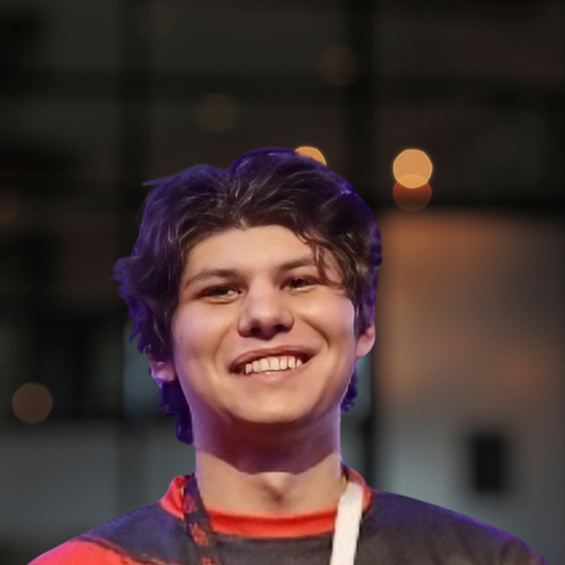 A young person with wavy dark hair smiles warmly, wearing a red and black Bitcube shirt with a white lanyard. The background is softly blurred, aglow with warm, out-of-focus lights.