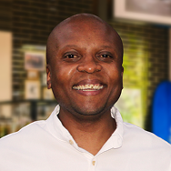 A man wearing a white shirt is smiling at the camera. The softly blurred background, with green and brown tones, hints at Bitcube’s modern indoor decor.