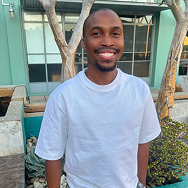 A smiling man in a white t-shirt stands outdoors in front of the modern Bitcube building, with large windows, trees, and plants visible in the background.