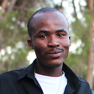 A man with short hair and a mustache wearing a black Bitcube shirt over a white t-shirt stands outdoors, with blurred trees in the background.