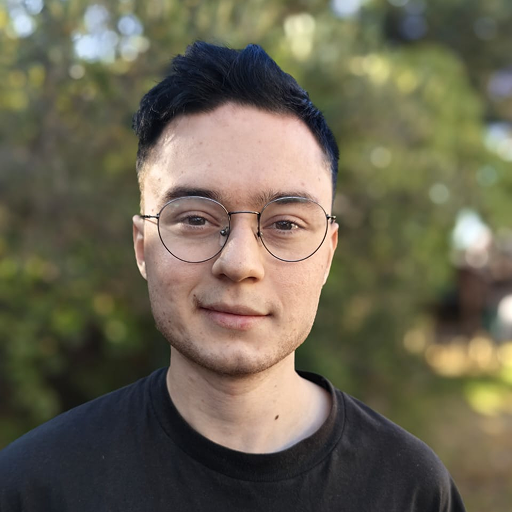 A person with short dark hair and round glasses is smiling slightly, wearing a dark Bitcube shirt and standing outdoors with green trees and sunlight in the blurred background.